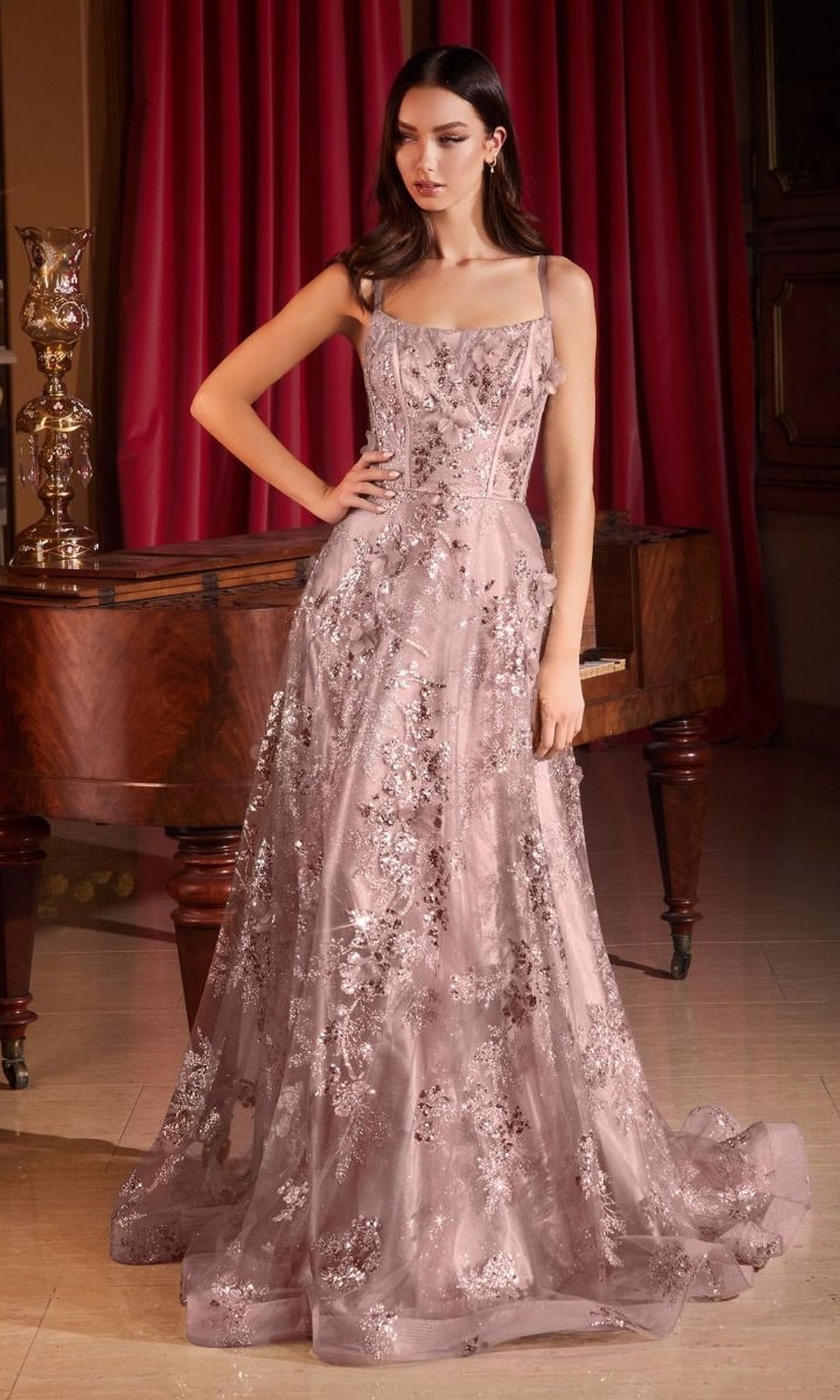 A woman wearing the Long Formal Dress CB144 by Ladivine, a floor-length sleeveless pink gown with floral embroidery and subtle glitter embellishments, stands in front of a piano and red curtains.